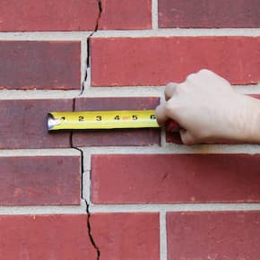 Hand holding a measuring tape against a cracked brick wall, showing the size of the crack in the masonry